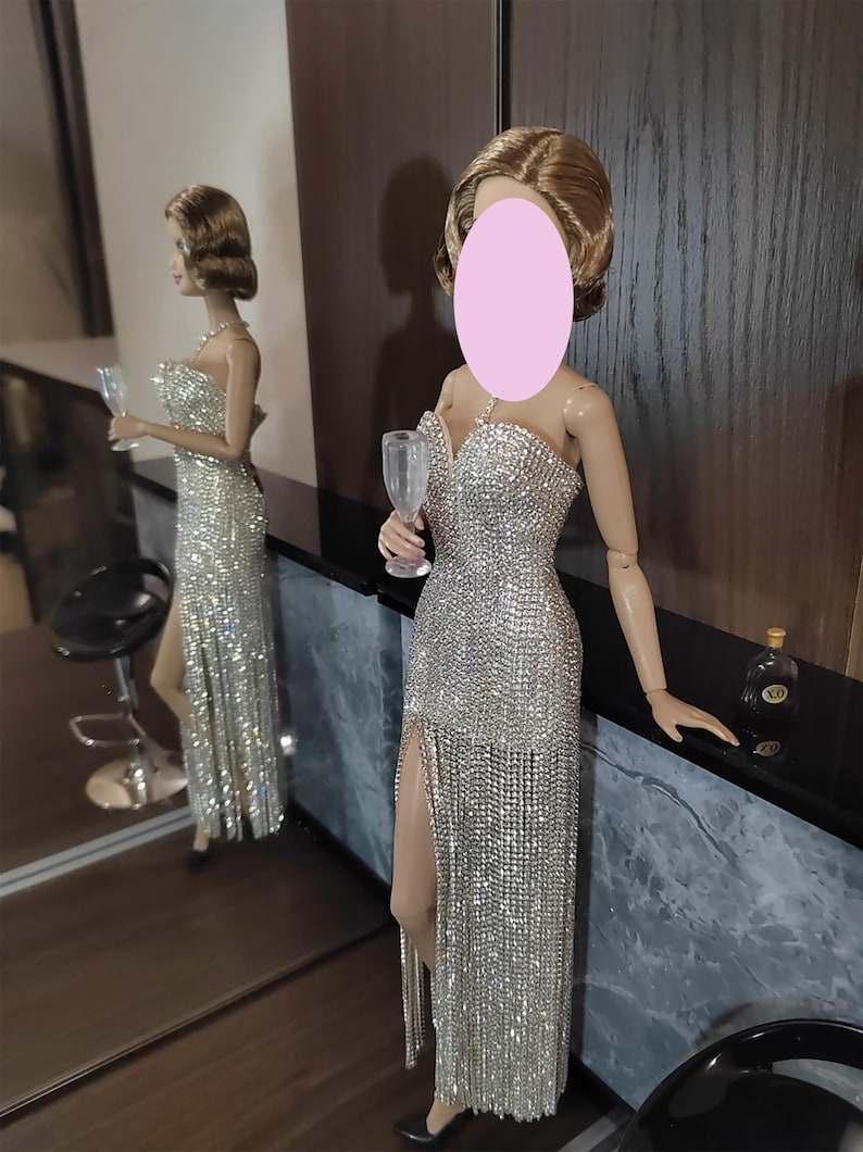 May include: A doll in a dazzling silver evening gown with a high split and a champagne glass. The dress is adorned with shimmering details. The doll is positioned near a mirror, reflecting the gown and the surroundings.