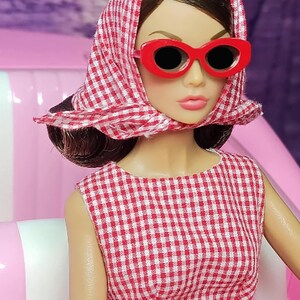K&ouml;nnte beinhalten: Eine Puppe in einem rot-wei&szlig; karierten Kleid mit rotem G&uuml;rtel und rotem Kopftuch. Die Puppe tr&auml;gt eine rote Sonnenbrille und hat braunes Haar.