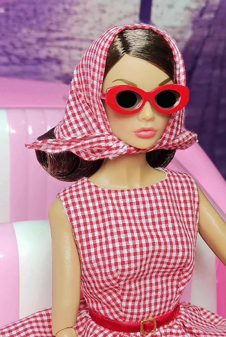 K&ouml;nnte beinhalten: Eine Puppe in einem rot-wei&szlig; karierten Kleid mit rotem G&uuml;rtel und rotem Kopftuch. Die Puppe tr&auml;gt eine rote Sonnenbrille und hat braunes Haar.