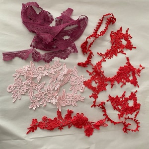 May include: Assortment of lace trims in pink, red, and burgundy. The lace trims are delicate and intricate, with floral patterns.