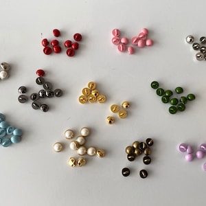 Op de afbeelding: Assortiment van kleine, ronde knopen in verschillende kleuren, waaronder rood, roze, blauw, groen, geel, goud, zilver en wit. De knopen hebben een platte achterkant en een verhoogde koepel aan de voorkant.