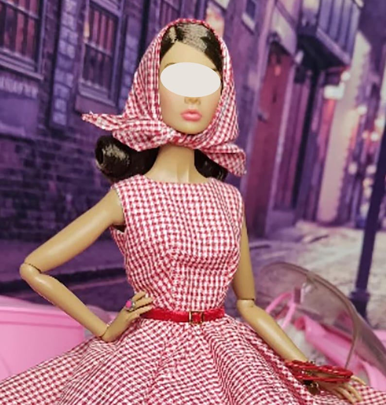 K&ouml;nnte beinhalten: Eine Puppe in einem rot-wei&szlig; karierten Kleid mit passendem Kopftuch. Die Puppe tr&auml;gt einen roten G&uuml;rtel und h&auml;lt eine kleine Handtasche.