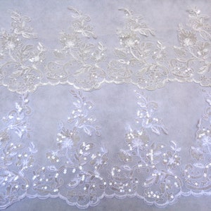 May include: Close-up of white lace fabric with floral and sequin embellishments. The lace features a scalloped edge and intricate designs of flowers and leaves. The fabric is ideal for bridal wear or other special occasion garments.