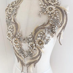 Può includere: Un'applicazione per corpetto ornata e scintillante con un design floreale. L'applicazione è riccamente impreziosita da cristalli trasparenti, perline e paillettes, creando un effetto abbagliante. Il design è posto su un tessuto trasparente bianco sporco, perfetto per abiti da sposa o da sera.
