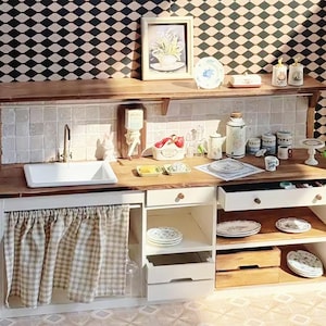 Op de afbeelding: Een miniatuurkeuken met een houten aanrecht, witte kasten en een geruit gordijn. De scène omvat een gootsteen, borden en decoratieve items zoals een ingelijste foto en kleine potjes. De algehele esthetiek is rustiek en charmant.
