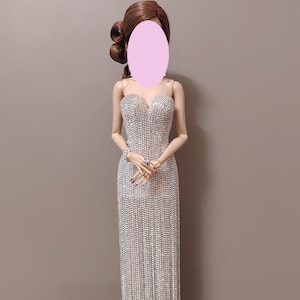 May include: A doll in a silver, floor-length gown. The strapless dress is adorned with sparkling embellishments. The doll has brown hair styled in curls. The background is a plain, neutral colour.
