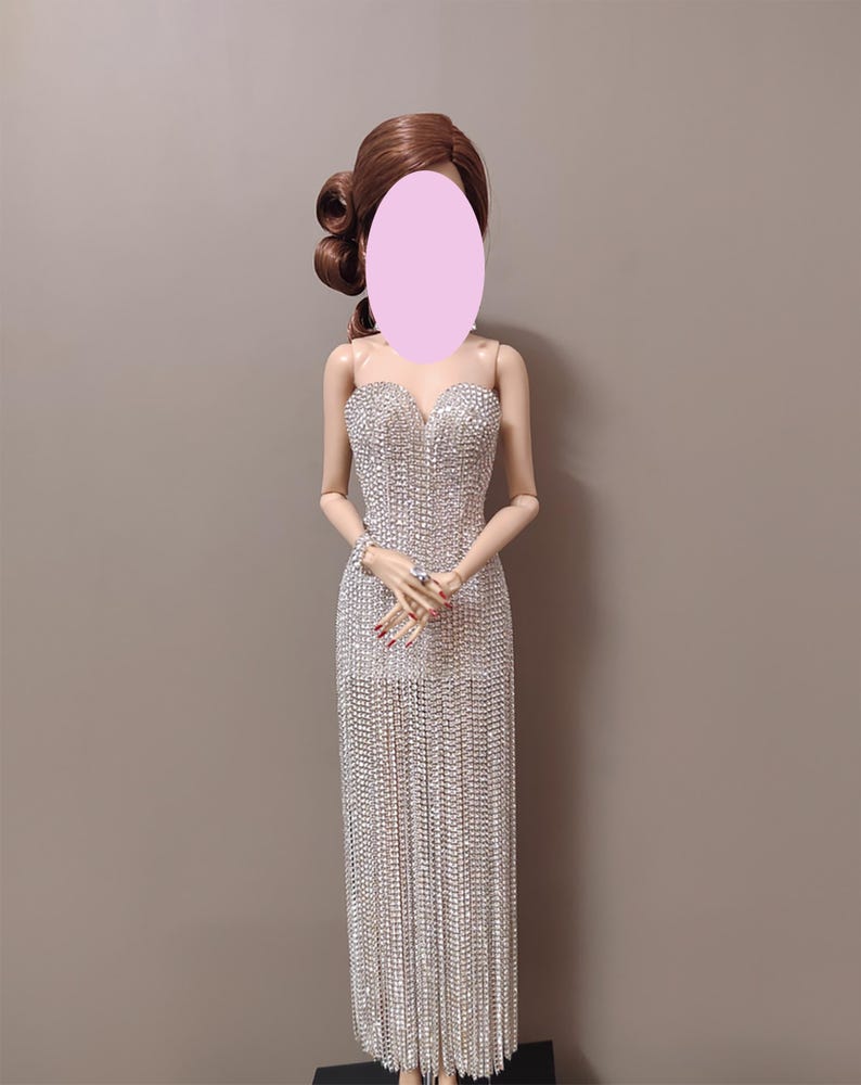 May include: A doll in a silver, floor-length gown. The strapless dress is adorned with sparkling embellishments. The doll has brown hair styled in curls. The background is a plain, neutral colour.