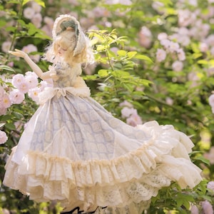 Puede incluir: Mu&ntilde;eca con un vestido de estilo vintage con un estampado azul claro y crema, adornado con encaje y volantes. La mu&ntilde;eca tiene el pelo rubio y lleva un gorro a juego. El fondo presenta flores rosas y vegetaci&oacute;n.