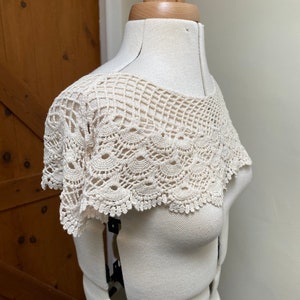 May include: A white crocheted collar with a delicate lace pattern. The collar is displayed on a white dress form.