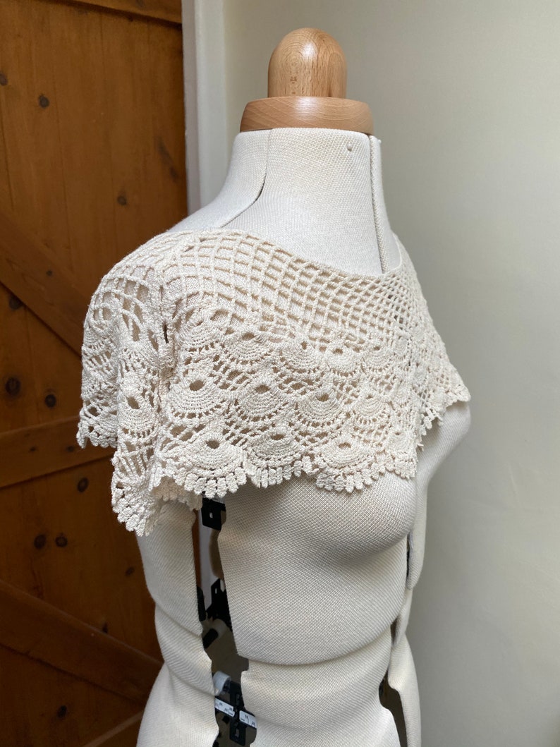 May include: A white crocheted collar with a delicate lace pattern. The collar is displayed on a white dress form.