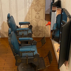 K&ouml;nnte beinhalten: Zwei Vintage-Friseurst&uuml;hle mit blauer Polsterung und Metallrahmen. Die St&uuml;hle stehen in einem Raum mit einem Spiegel und Holzboden.