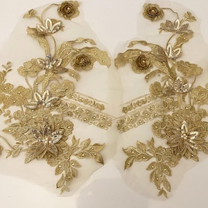May include: Two gold floral appliques with detailed embroidery, sequins, and bead embellishments. The appliques feature flowers, leaves, and decorative borders, set against a sheer, off-white background. The design is symmetrical, with each applique mirroring the other.