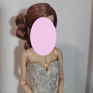 May include: A doll with auburn hair styled in an updo. The doll is dressed in a shimmering silver strapless gown and a matching bracelet. A delicate silver necklace with a pendant is around the doll's neck. The doll's face is obscured by a pink oval.