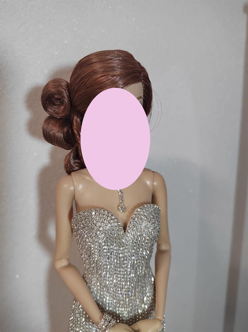 May include: A doll with auburn hair styled in an updo. The doll is dressed in a shimmering silver strapless gown and a matching bracelet. A delicate silver necklace with a pendant is around the doll's neck. The doll's face is obscured by a pink oval.