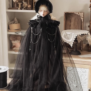 May include: A doll with pale skin and blonde hair, dressed in a black bonnet and a long, flowing black tulle cape. The cape features a beaded chain and a cross pendant. The doll is positioned before a white shelf.