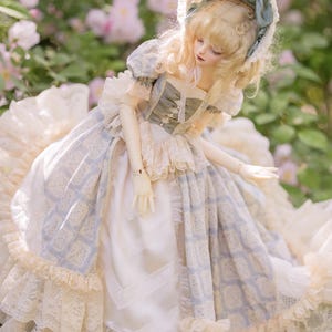 Puede incluir: Una mu&ntilde;eca con un vestido y un gorro de estilo vintage. El vestido combina telas azul claro y blanco, adornado con encaje y patrones. La mu&ntilde;eca tiene el pelo rubio y posa en un jard&iacute;n con flores rosas.