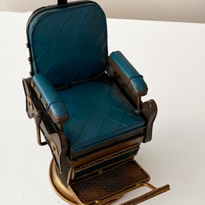 K&ouml;nnte beinhalten: Ein blauer Barbersitz aus Metall mit goldener Basis und Fu&szlig;st&uuml;tze. Der Stuhl hat eine gepolsterte Sitzfl&auml;che und R&uuml;ckenlehne sowie eine Kopfst&uuml;tze. Der Stuhl ist ein Dekorationsst&uuml;ck.