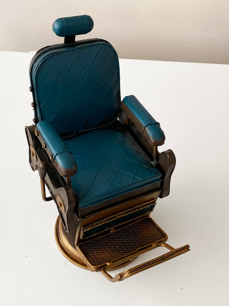 K&ouml;nnte beinhalten: Ein blauer Barbersitz aus Metall mit goldener Basis und Fu&szlig;st&uuml;tze. Der Stuhl hat eine gepolsterte Sitzfl&auml;che und R&uuml;ckenlehne sowie eine Kopfst&uuml;tze. Der Stuhl ist ein Dekorationsst&uuml;ck.