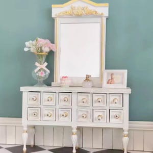 Könnte beinhalten: Ein weißer Schminktisch mit Spiegel, verziert mit goldenen Akzenten. Der Schminktisch hat mehrere Schubladen mit goldenen Knöpfen. Eine Blumenvase, ein kleiner Kuchen, eine Parfümflasche und ein gerahmtes Foto sind ausgestellt.