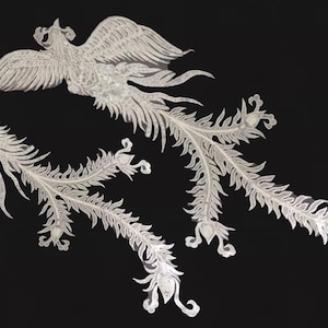 May include: Two detailed, white phoenix designs with outstretched wings and flowing tail feathers against a black background. The birds are intricately crafted, showcasing a delicate, lacy appearance, possibly for decorative use.