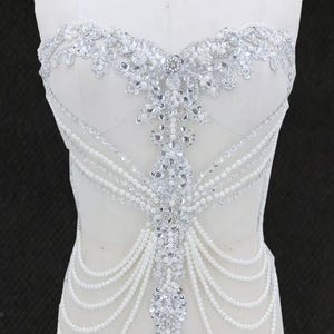 May include: A white bodice adorned with silver beading and pearl detailing. The design includes a strapless neckline, a central beaded motif, and flowing pearl strands, offering a glamorous and elegant aesthetic.
