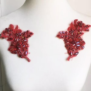 Può includere: Due applicazioni floreali rosse con intricate perline e dettagli di paillettes. Le applicazioni sono realizzate con filo rosso e presentano una varietà di perline rosse e iridescenti. Sono progettate per essere cucite su tessuto.
