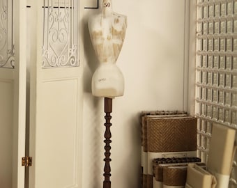 May include: A vintage-style dressmaker's mannequin with a cream-colored torso and a wooden stand. The mannequin is labeled "50" at the neck and "DOLL" at the base. Rolls of fabric are stacked nearby, suggesting a sewing or design theme.