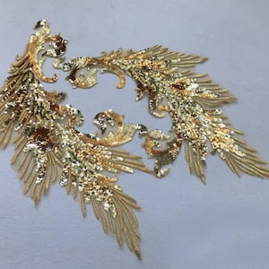 Può includere: Due applicazioni ricamate dorate e ornate. Ogni applicazione presenta un design a piuma stilizzato con dettagli intricati e paillettes scintillanti. Le applicazioni sono ideali per abbellire capi di abbigliamento o progetti artigianali.