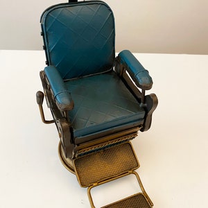 K&ouml;nnte beinhalten: Ein blauer und goldener Metall-Friseurstuhl mit Fu&szlig;st&uuml;tze. Der Stuhl hat einen gepolsterten Sitz und R&uuml;ckenlehne, und die Fu&szlig;st&uuml;tze ist verstellbar.