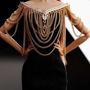 May include: A black dress with a gold chain necklace and matching shoulder piece. The chain is made of multiple strands of different sizes and has a large, clear crystal pendant.