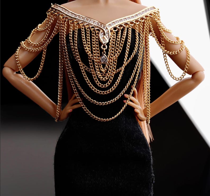May include: A black dress with a gold chain necklace and matching shoulder piece. The chain is made of multiple strands of different sizes and has a large, clear crystal pendant.