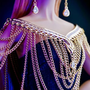 May include: A gold chain necklace with a decorative design and dangling crystal accents. The necklace is worn by a doll with long blonde hair.