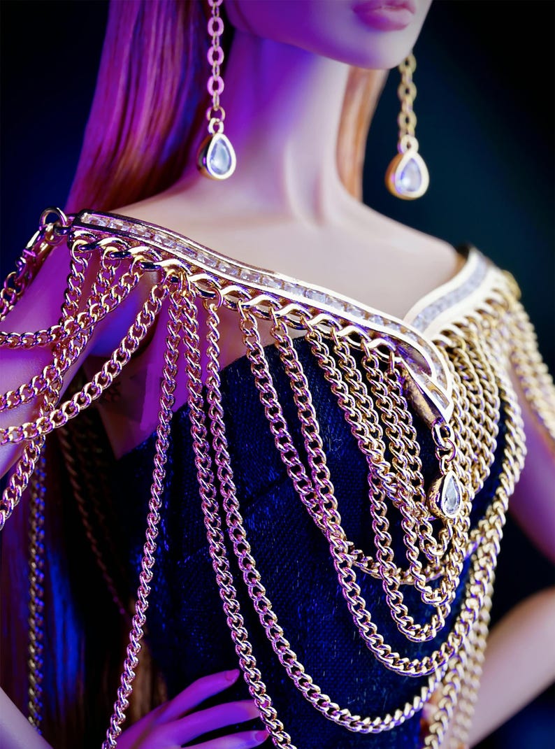 May include: A gold chain necklace with a decorative design and dangling crystal accents. The necklace is worn by a doll with long blonde hair.