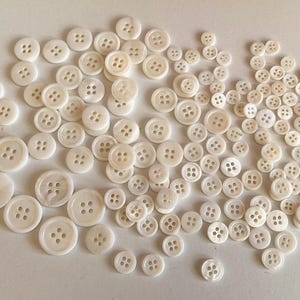 May include: A large collection of white mother of pearl buttons of various sizes. The buttons are arranged in a random pattern on a white surface.