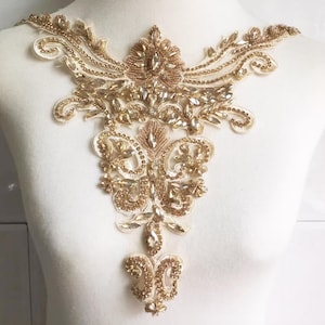 以下が含まれることがあります： 金とクリーム色のビーズ刺繍アップリケ。精巧な花柄のデザインが特徴です。キラキラ光るラインストーンとシードビーズで装飾され、豪華な印象を与えます。衣類やアクセサリーの装飾に最適です。
