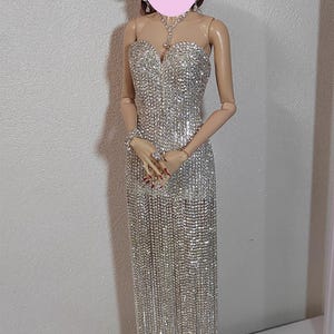 May include: A doll in a shimmering silver evening gown with a sweetheart neckline and a flowing skirt. The doll has brown hair styled in an updo and is wearing a matching necklace, earrings, and bracelet. The doll is standing on a black base.