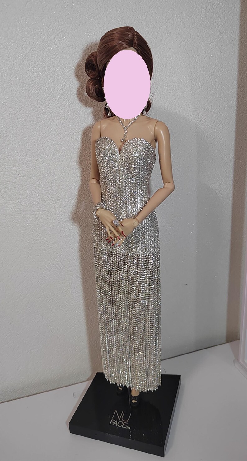 May include: A doll in a shimmering silver evening gown with a sweetheart neckline and a flowing skirt. The doll has brown hair styled in an updo and is wearing a matching necklace, earrings, and bracelet. The doll is standing on a black base.