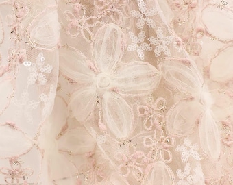 May include: Close-up of a delicate fabric featuring a floral design. The fabric is a soft, off-white color with layers of sheer material. Intricate, three-dimensional flowers in white and pale pink are scattered across the surface, creating a textured effect.