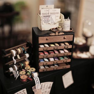 Puede incluir: Exhibición en miniatura de suministros de artesanía con un mueble de madera con cajones llenos de muestras de tela de colores. Bobinas de hilo y encaje dispuestas cerca. Un pequeño portacarteles muestra tarjetas impresas.