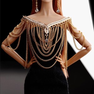 May include: A gold chain necklace and matching shoulder piece with clear rhinestones. The necklace and shoulder piece are worn by a doll with red hair and a black dress.