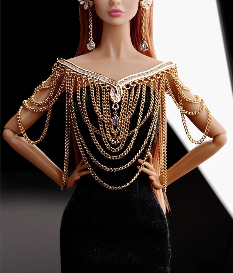 May include: A gold chain necklace and matching shoulder piece with clear rhinestones. The necklace and shoulder piece are worn by a doll with red hair and a black dress.