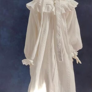 May include: A white cotton nightgown with a lace trim and a ruffled collar. The nightgown has long sleeves and a button-up front.