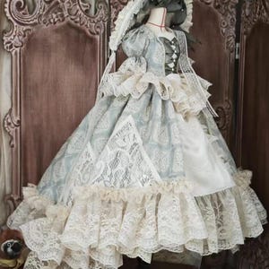 Puede incluir: Vestido de mu&ntilde;eca de estilo vintage con un estampado floral azul claro y blanco, adornado con capas de delicado encaje. El vestido presenta un cuello alto, mangas abullonadas y un gorro a juego. El vestido se exhibe en un maniqu&iacute;.