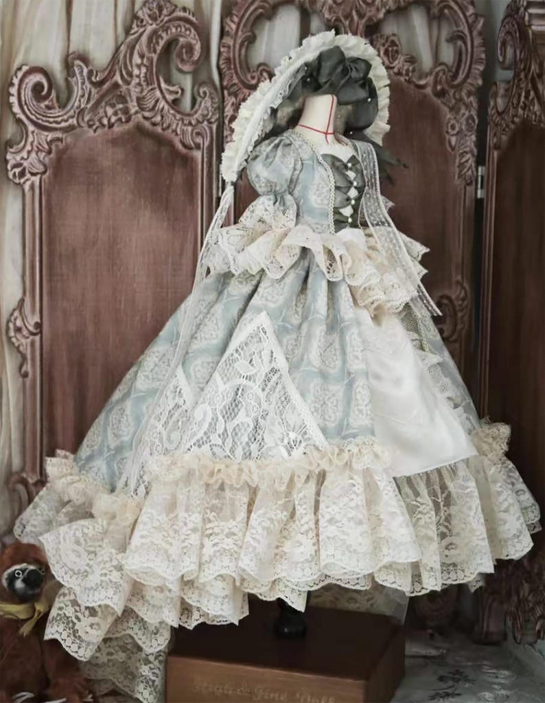 Puede incluir: Vestido de mu&ntilde;eca de estilo vintage con un estampado floral azul claro y blanco, adornado con capas de delicado encaje. El vestido presenta un cuello alto, mangas abullonadas y un gorro a juego. El vestido se exhibe en un maniqu&iacute;.