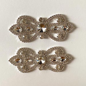 Könnte beinhalten: Zwei silberne Strass-Verzierungen in Form einer Schleife. Jede Schleife besteht aus kleinen, klaren Strasssteinen und hat einen größeren, klaren Strassstein in der Mitte.