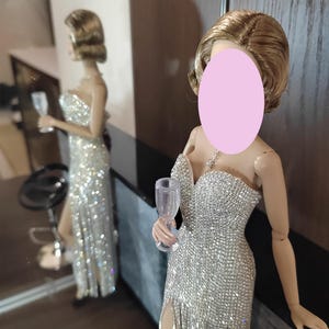 May include: A doll in a dazzling silver gown with a high slit, holding a clear wine glass. The dress is covered in shimmering embellishments. The doll's hair is styled in a short, wavy bob. A mirror reflects the doll and the room's interior.