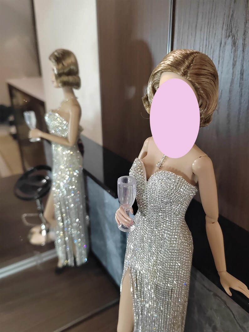 May include: A doll in a dazzling silver gown with a high slit, holding a clear wine glass. The dress is covered in shimmering embellishments. The doll's hair is styled in a short, wavy bob. A mirror reflects the doll and the room's interior.