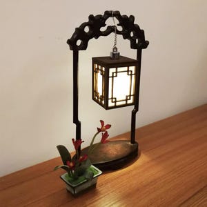Könnte beinhalten: Eine dekorative, dunkelbraune Laterne mit quadratischem, beleuchtetem Schirm. Die Laterne hängt an einem dekorativen Bogen und wird von zwei vertikalen Pfosten getragen. Am Sockel befindet sich eine kleine Topfpflanze mit roten Blüten.