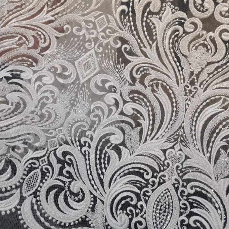 Puede incluir: Primer plano de tela de encaje blanco con un intrincado dise&ntilde;o floral y de volutas. El encaje presenta patrones en relieve y texturizados y acentos brillantes, creando una apariencia delicada y elegante. El dise&ntilde;o incluye formas de diamante y motivos florales detallados.