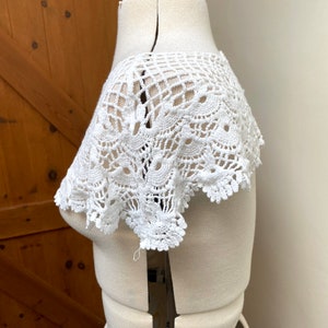 May include: White crochet lace collar with a delicate floral pattern. The collar is displayed on a white dress form.